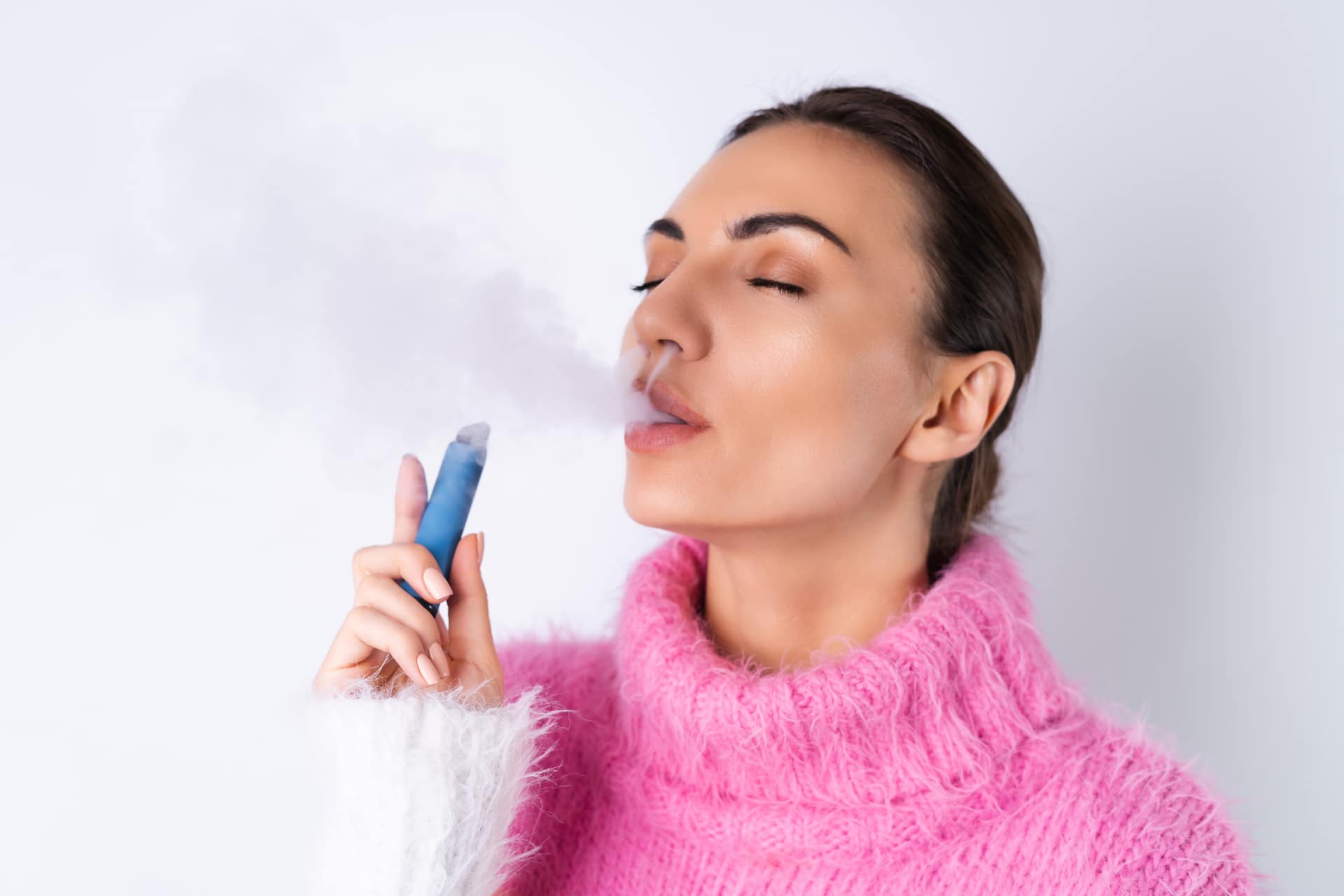 Person in a pink sweater vaping with closed eyes, holding a blue e-cigarette against a plain white background. Berry Good Vibes E-Liquid.